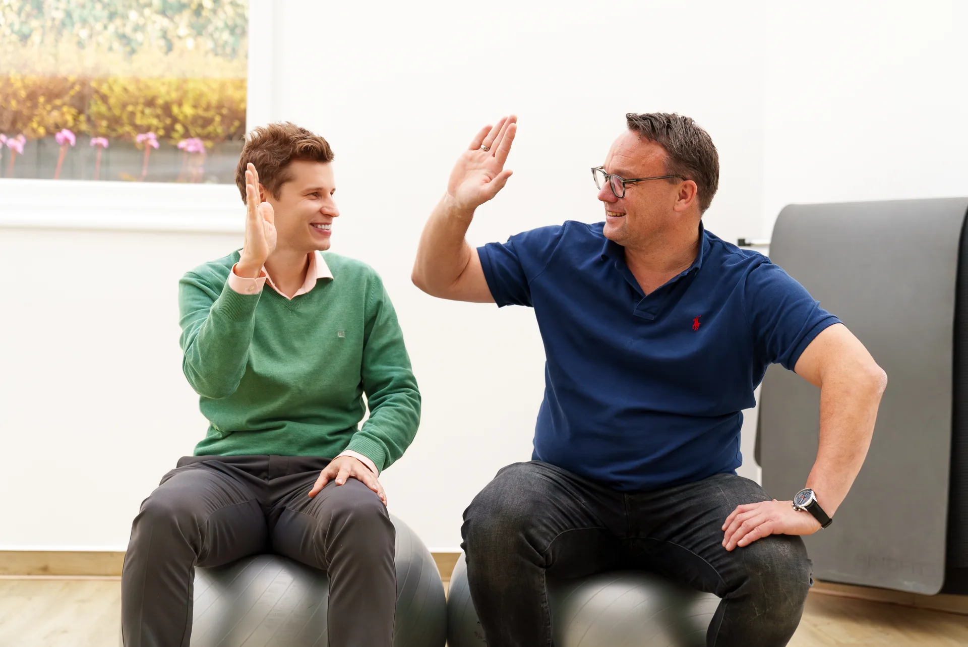 Zwei lachende Mitarbeiter geben sich ein High-Five auf Gymnastikbällen beim Dreh für ein YouTube-Recruiting-Video für neue Physiotherapeut-Jobs bei PHYSIOMED SÜD in Berlin.
